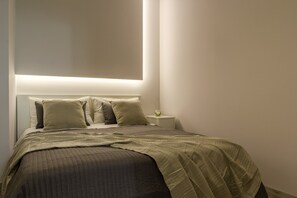 Ein ordentlich bezogenes Bett mit einer gesteppten Decke, ein Nachttisch mit einer Lampe und eine Wandleuchte.