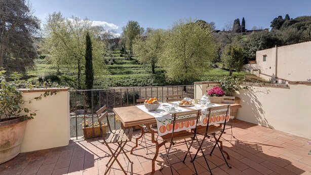 Un patio exterior con una mesa dispuesta para comer, rodeado de plantas en macetas y vistas a un jardín en terrazas.