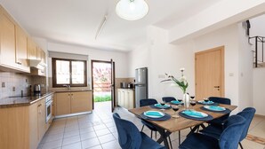 Et moderne køkken med træskabe, en indbygget ovn og et køleskab. Der er et spiseområde med et bord dækket til fire personer, blå stole og en vase med blomster.