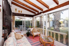 A sunlit patio with wicker furniture, a glass door leading outside, and a view of a garden.