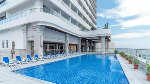 Piscina com espreguiçadeiras, edifício com varandas e plantas em vasos.