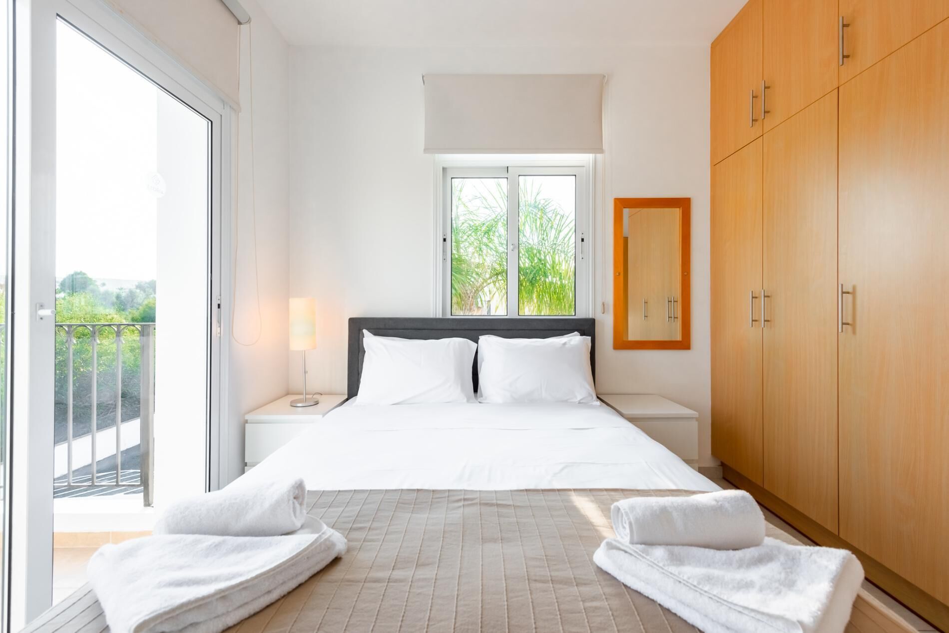 Een hotelkamer met een bed, een nachtkastje, een lamp, een raam met uitzicht op groen en een houten garderobe.