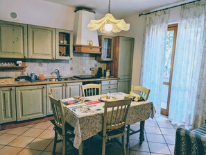 Une cuisine avec une table à manger, des chaises, un lustre et une vue sur l’extérieur à travers une fenêtre ornée de rideaux.