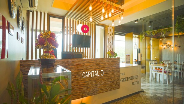 A reception desk with a wooden finish and the name "CAPITAL O" prominently displayed. There is a television mounted on the wall above the desk, and a large window providing natural light. The reception area is adjacent to a dining area with white chairs and a table. - Capital O Vaccinated Staff - OYO 785 The Greenhive (Ibaan)