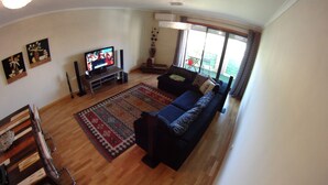 A living room with a dark blue sofa, a flat-screen TV on a stand, a patterned rug, and artwork on the walls. - Quinta do Anjo Apartment (Palmela)