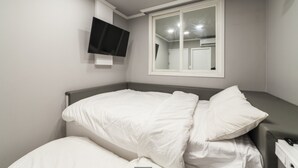 Un lit bien fait, avec une literie blanche, dans une pièce où l’on aperçoit un téléviseur fixé au mur et une fenêtre.