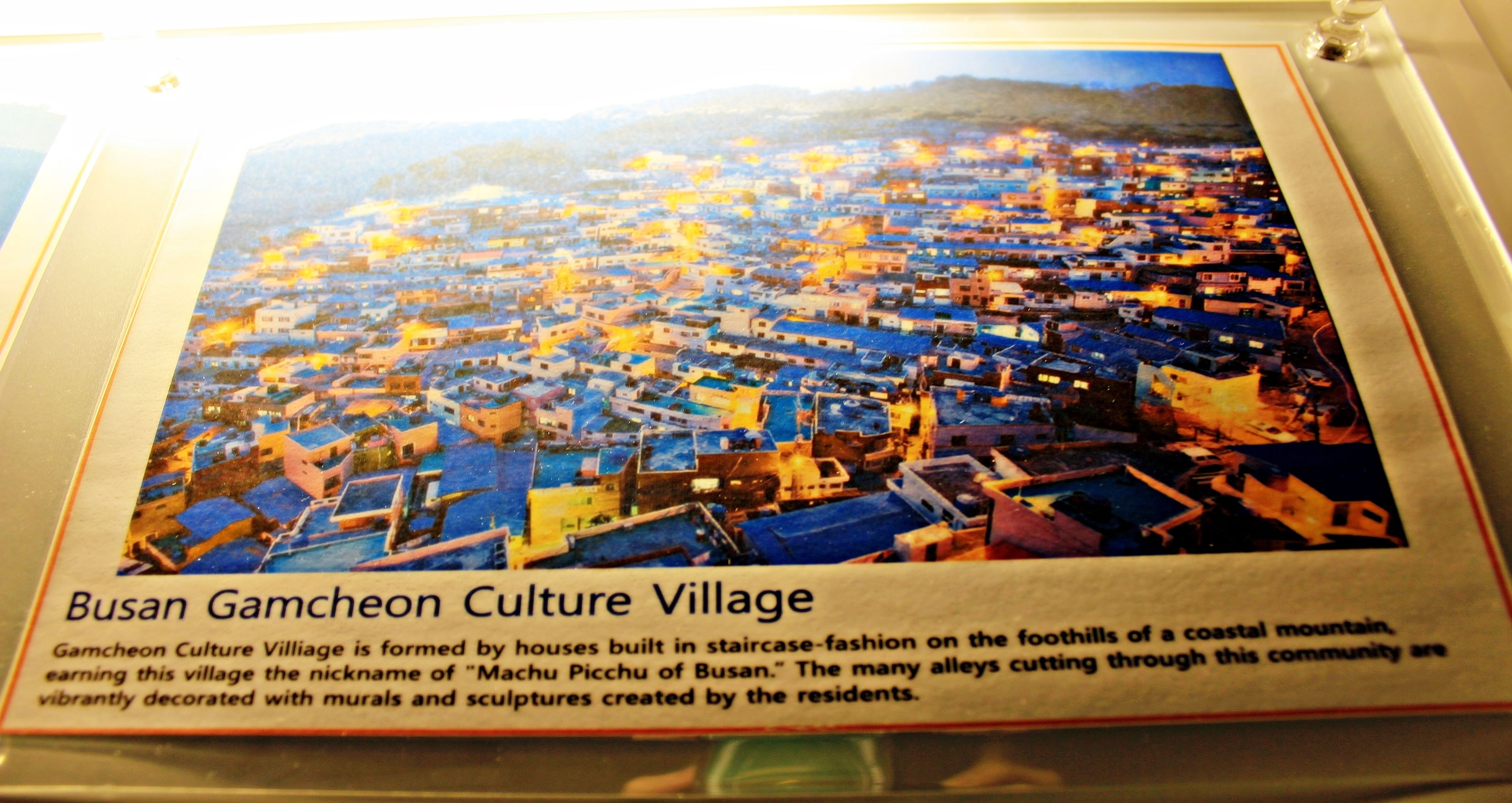 A framed picture of Busan Gamcheon Culture Village, featuring houses built in staircase-fashion on the foothills of a coastal mountain, earning the nickname "Machu Picchu of Busan." The many alleys cutting through this community are vibrantly decorated with murals and sculptures created by the residents.