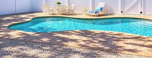 Una piscina con cerca blanca, muebles de patio y palmeras al fondo.
