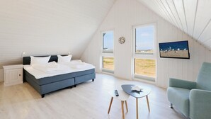 Ein Schlafzimmer mit Bett, Nachttisch, Sessel, kleinem Tisch und Fernseher. Fenster bieten Blick auf den Strand.