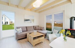 Een moderne woonkamer met een beige bank, een houten salontafel en een lichtblauwe fauteuil. Er is een groot raam met uitzicht op een huis en een grasveld.
