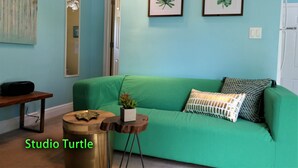 Een kamer met teal muren, een groene bank, een kleine houten tafel en een rond krukje. Twee ingelijste botanische prints hangen aan de muur.
