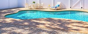 A swimming pool with a brick patio, white outdoor furniture, and a white fence.