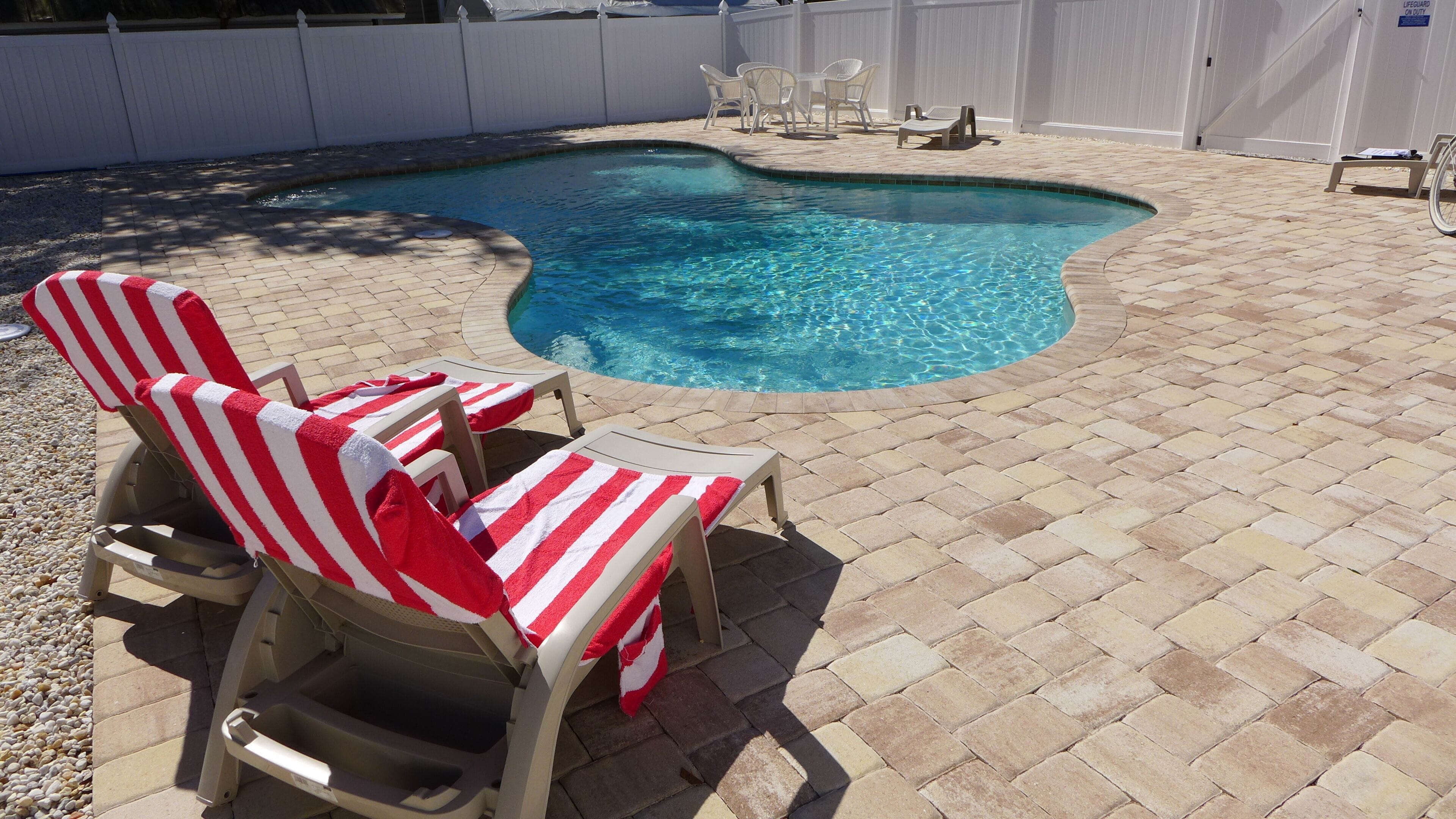 A swimming pool with a brick patio, lounge chairs with striped towels, and a white fence in the background.