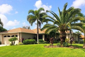 Una casa residencial con césped cuidado, palmeras y un cielo azul despejado.