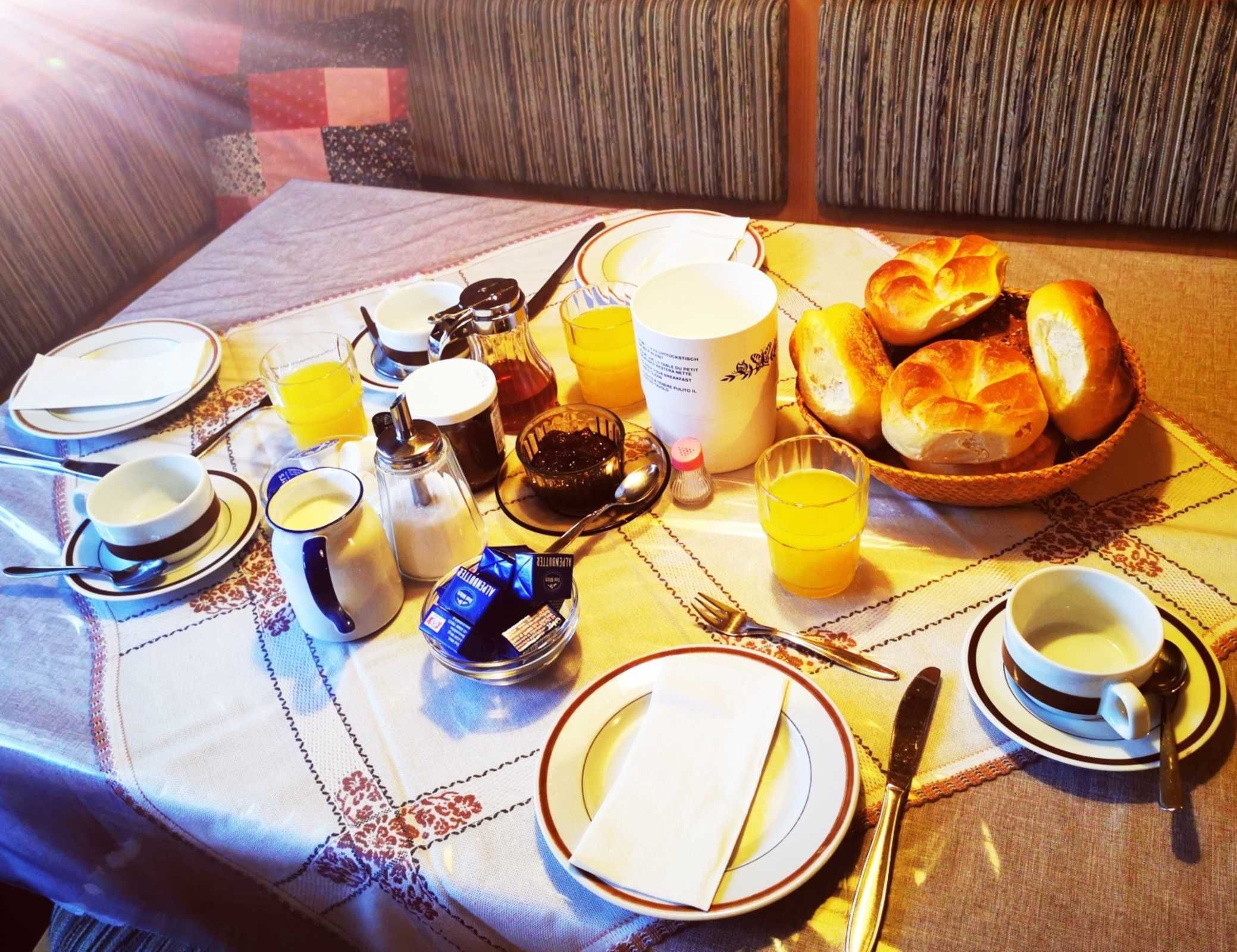 Une table de petit-déjeuner dressée avec des assiettes, des tasses, un panier de pain, une cafetière et une bouteille de jus de fruits.