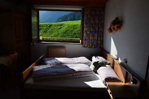 Una cama con colcha a cuadros, una ventana con vistas a colinas verdes y una cortina con estampado floral.