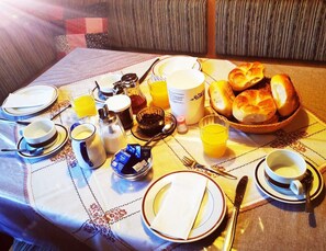 Ein Frühstückstisch reichhaltig gedeckt mit Tellern, Tassen, einem Brotkorb, einer Kaffeekanne und verschiedenen Gewürzen.