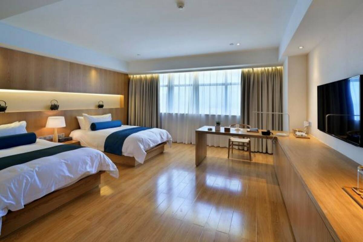A hotel room with two beds, a desk, a TV, and a large window with curtains.
