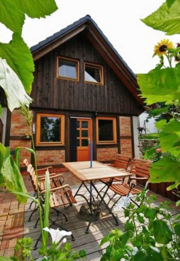 Une cabane en bois avec un toit en pente, une porte d’entrée et deux fenêtres. Devant la cabane, on trouve une terrasse en bois avec une table et des chaises.