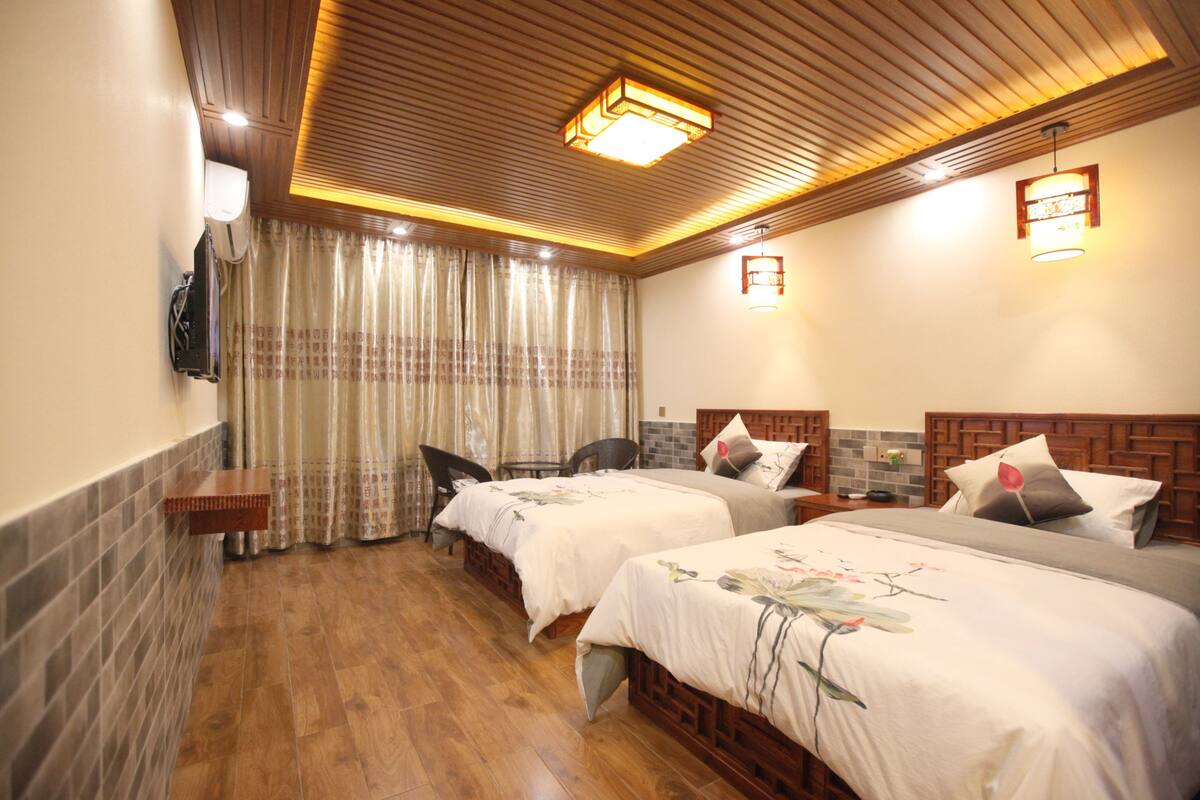 A hotel room with two beds, a wooden ceiling, a wall-mounted air conditioner, and a window with curtains.