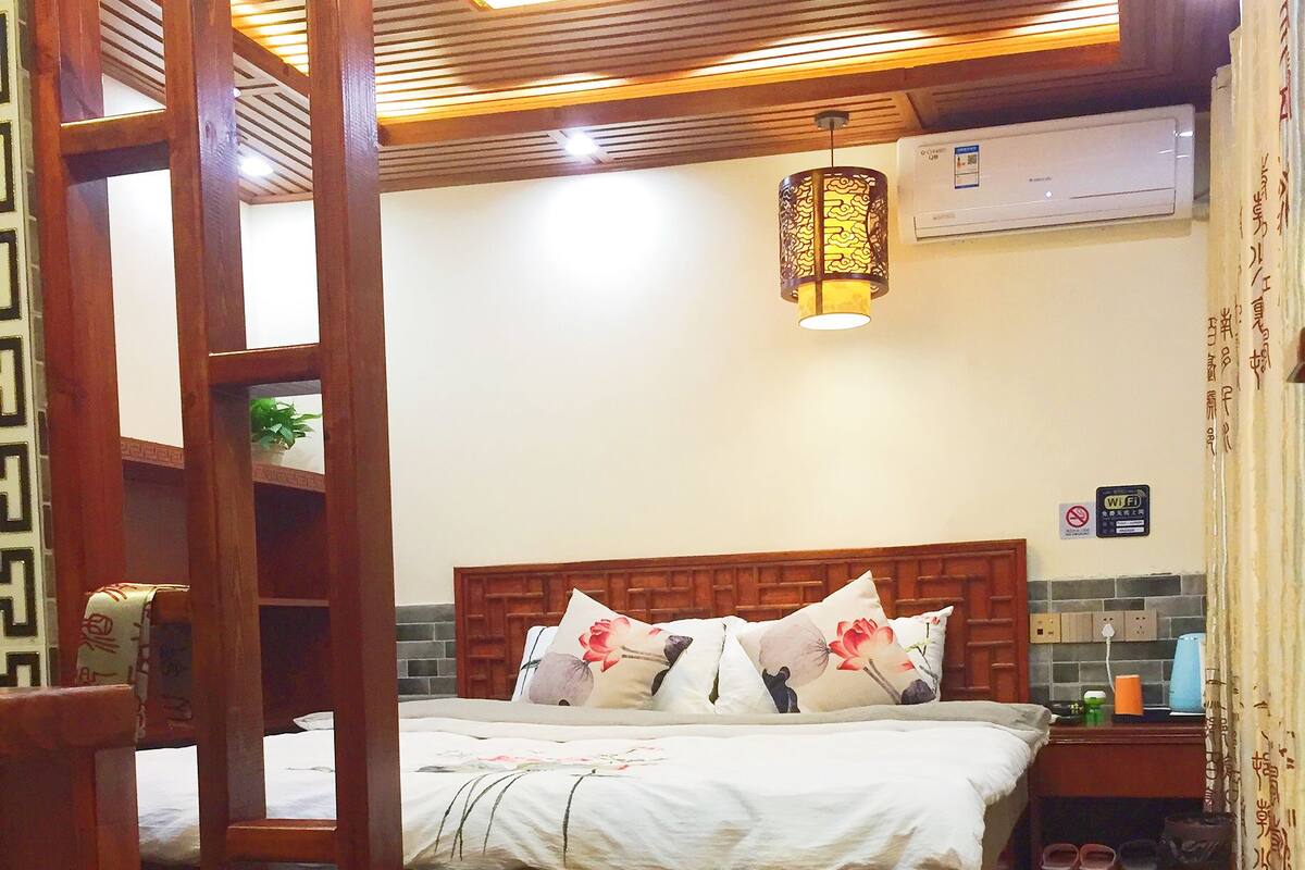 A bedroom with a wooden bed frame, white bedding, and a wooden ladder. There is a wall-mounted air conditioner and a small table with a lamp.