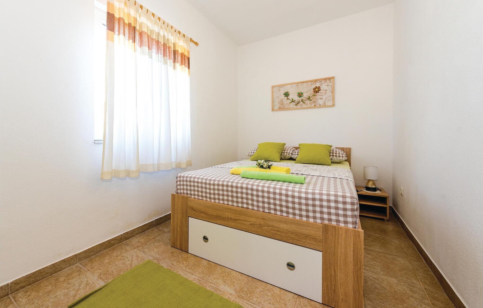 Un dormitorio con colcha a cuadros, almohadas verdes, una mesita de noche con lámpara y una ventana con cortinas translúcidas.