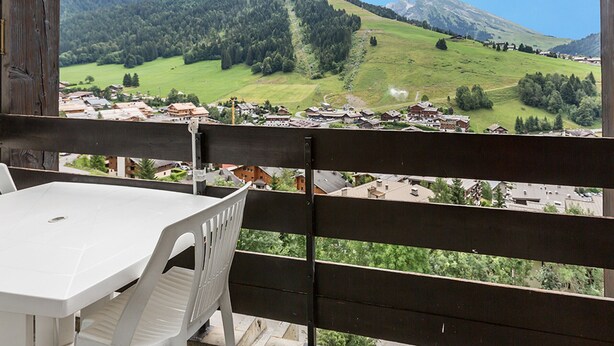 A balcony with a white table and chairs, overlooking a village with green fields and mountains in the background. - South-Facing Studio Near Aquatic Area, Pets Allowed (La Clusaz)
