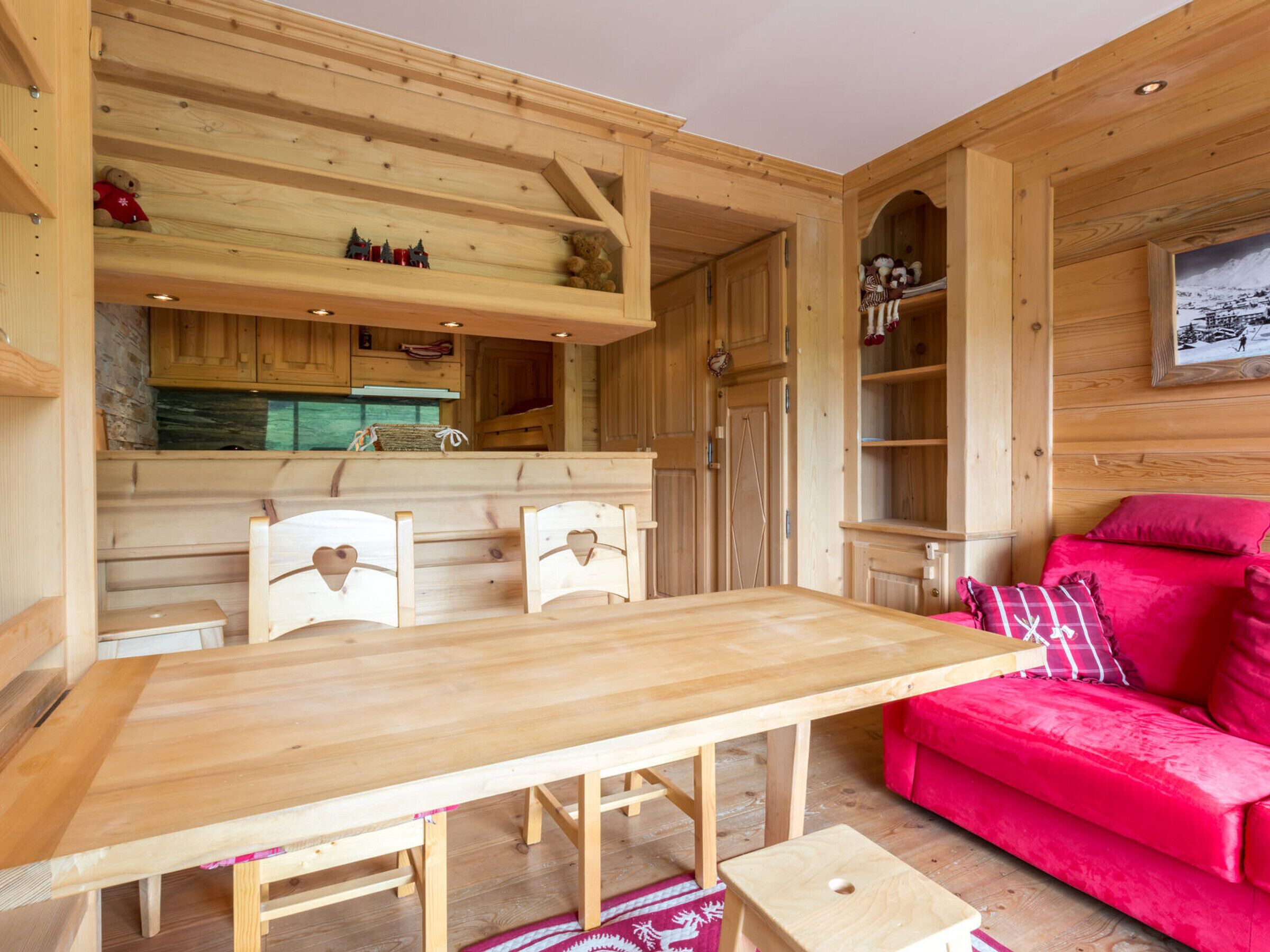 A wooden interior with a dining table, two chairs, a red sofa, and a kitchen area with cabinets and a clock.