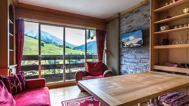 A cozy room with a large window, a wooden table, a red sofa, and a TV mounted on a stone wall.
