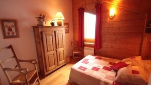 A cozy bedroom with a wooden bed, a chair, a cabinet, and a window with curtains. - Stunning 4-Room Mezzanine with Parking and Pets Allowed (Villard-sur-Doron)