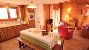 A cozy cabin-style room with a wooden interior, a dining table with a green tablecloth, a bench, a red armchair, and a television. - Stunning 4-Room Mezzanine with Parking and Pets Allowed (Villard-sur-Doron)