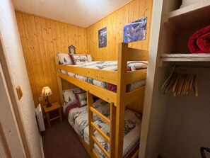 Et soveværelse med køjeseng, trævæg, et lille sengebord med lampe og en hylde med foldede klæder.