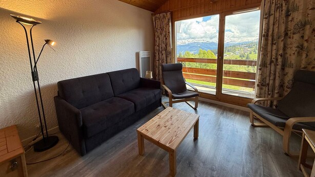 Habitación acogedora con techo de madera, un sofá oscuro, una mesa de centro de madera y una silla. Una lámpara de pie está a la izquierda, y una ventana con cortinas ofrece vistas a un paisaje montañoso.