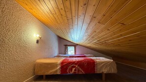 Un dormitorio con techo inclinado, una cama individual, un ventanal pequeño y una lámpara fijada a la pared.
