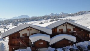 Una cabaña de madera con techo inclinado, rodeada de montañas y árboles cubiertos de nieve.