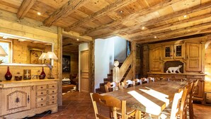 A rustic dining area with a long wooden table and chairs, a wooden cabinet with drawers, and a staircase leading to another room.