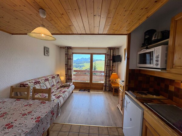 A cozy room with a wooden ceiling, a sofa with a floral cover, a small kitchen area with a microwave, and a view of mountains through a large window.