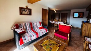 Una sala de estar acogedora con un sofá, un sillón rojo y una mesita de centro. Se distingue un área de comedor con una mesa y sillas al fondo.