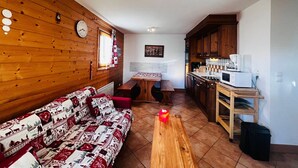 A cozy living room with a wooden wall, a sofa, a coffee table, and a kitchen area with wooden cabinets and a microwave.
