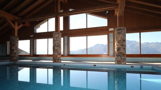 A large indoor swimming pool with stone pillars, wooden beams, and large windows offering mountain views.