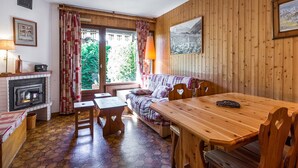 Een gezellige kamer met houten inrichting, een open haard, een bank, een eettafel en een groot raam met gordijnen.