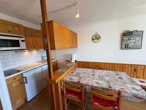Cozinha compacta com micro-ondas, pia e armários de madeira. Há uma mesa de jantar com cadeiras e um banco.