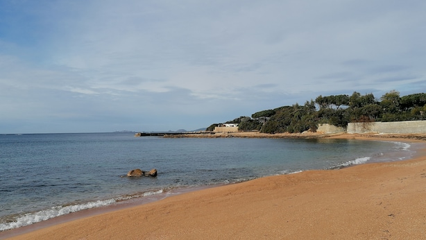 Ein Strand mit blauem Himmel, ruhigem Wasser und einem felsigen Ausläufer mit Vegetation.