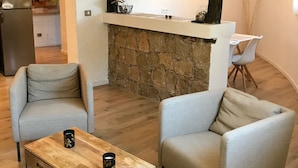Ein modernes Wohnzimmer mit zwei Sesseln, einem Couchtisch, einem Kamin aus Stein und einer Küche im Hintergrund.