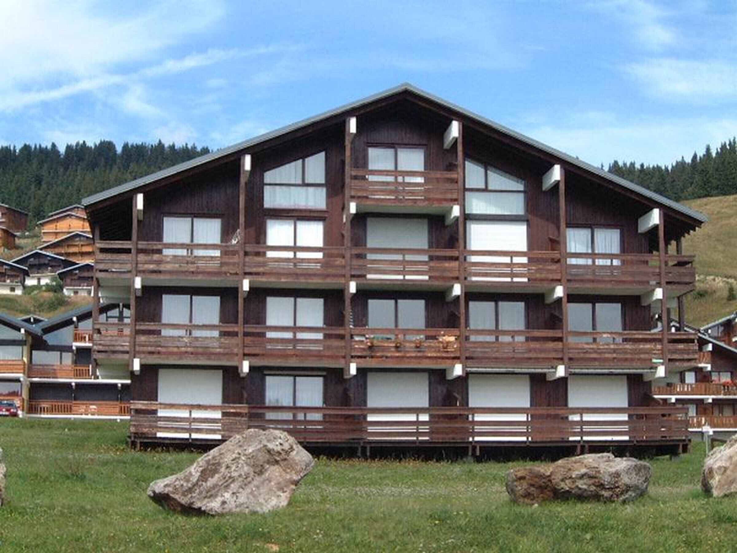 Un edificio de madera de varios pisos con balcones, ubicado en un área verde con rocas grandes y una colina boscosa al fondo.