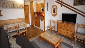 A cozy living room with a sofa, wooden coffee table, and a TV. There's a kitchen in the background with wooden cabinets and a microwave. - Studio cabin with large terrace and ideal location (Les Saisies)
