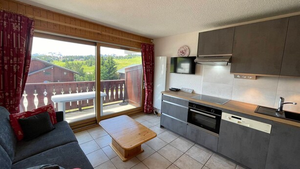 A modern kitchen with grey cabinets, a wooden countertop, and a built-in oven. There is a sliding glass door leading to a balcony with a view of a green landscape. A grey sofa and a wooden coffee table are in the living area.