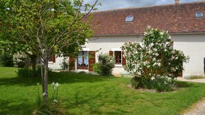 Una casa a due piani con tetto in tegole, un giardino con diverse piante e un piccolo albero con fiori bianchi.