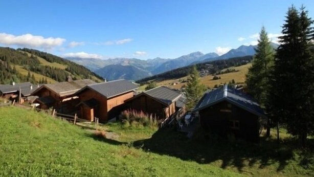 A row of wooden cabins with sloped roofs, set in a green valley with mountains in the background.