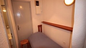 A narrow hallway with a door at the end, a small stool, and a bed with a wooden headboard.